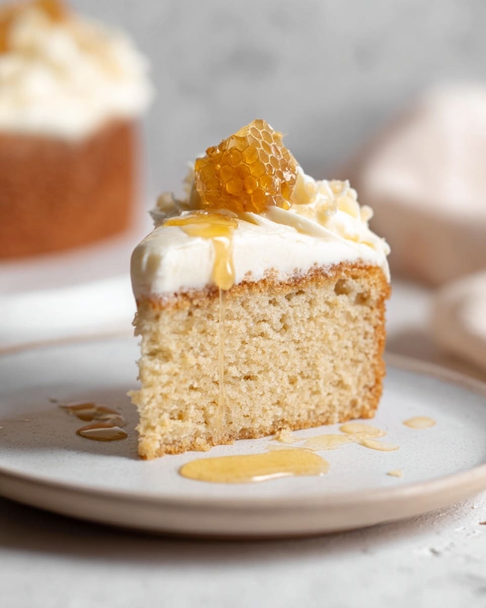 The image shows a single slice of cake on a white plate placed on a white marbled surface. The cake has two layers: a thick base layer that is light golden brown with a soft, spongy texture, and a creamy white frosting layer on top that looks smooth and fluffy. A small piece of honeycomb is placed on the center of the frosting, with honey drizzled over the top and dripping slightly down the sides of the cake and pooling lightly on the plate. The background is softly blurred with neutral tones. Photo taken with an iphone --ar 4:5 --v 7