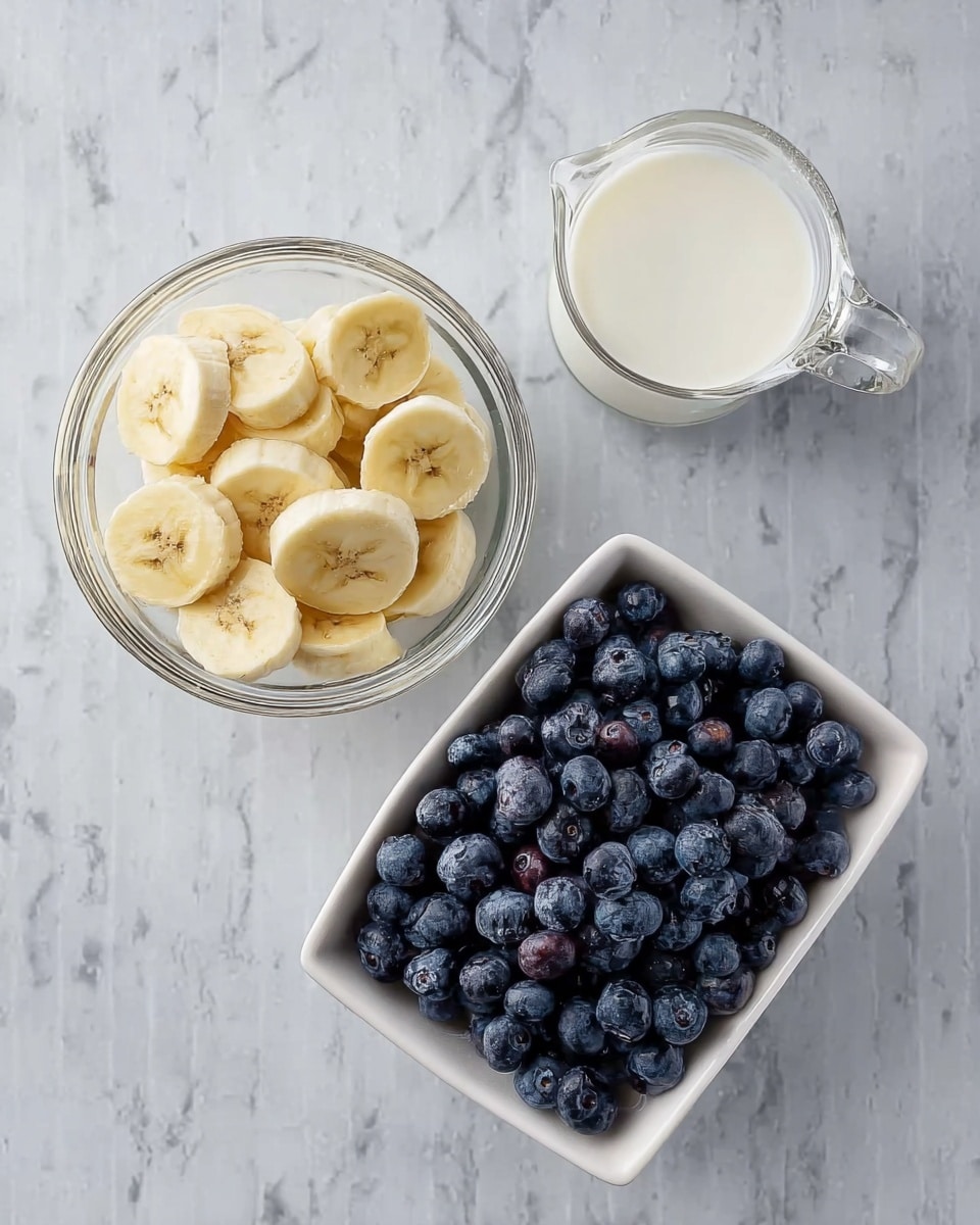 A small clear glass bowl filled with a layer of banana slices, showing creamy yellow and light brown colors, next to a small clear glass pitcher full of white liquid, and a white rectangular bowl full of fresh, plump blueberries with dark blue and purple tones. All items are placed on a white marbled surface. photo taken with an iphone --ar 4:5 --v 7