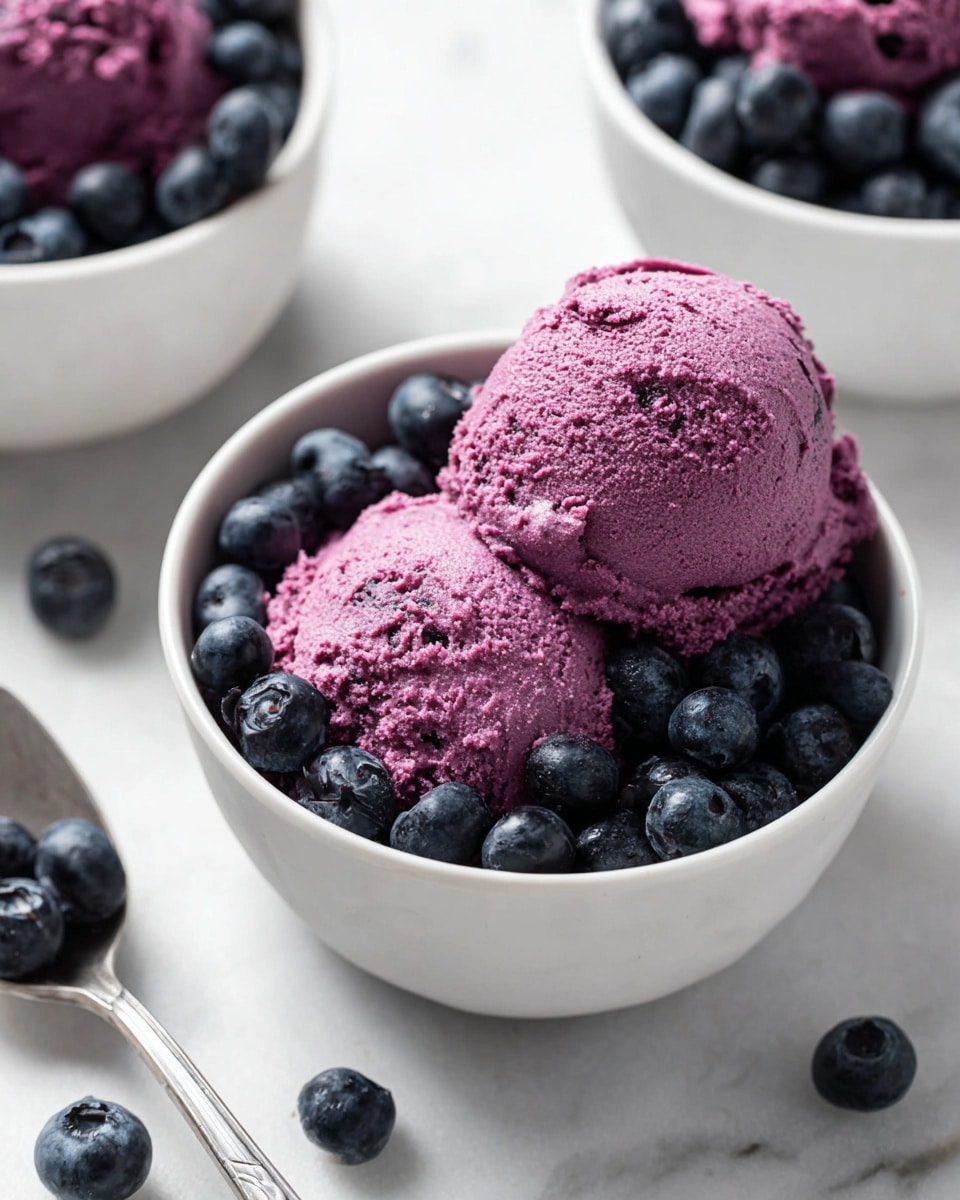 The image shows a white bowl filled with two scoops of thick, creamy purple blueberry mousse with a slightly textured surface. The mousse rests on a bed of whole, fresh blueberries, deep blue to almost black in color, filling about half the bowl at the bottom and peeking from the sides of the mousse. Around the main bowl, there are similar bowls with blueberries, all placed on a white marbled surface. Loose blueberries are scattered near the bowls, and a silver spoon holding two blueberries is visible in the lower left corner. The lighting is bright and natural, highlighting the colors and textures. Photo taken with an iphone --ar 4:5 --v 7