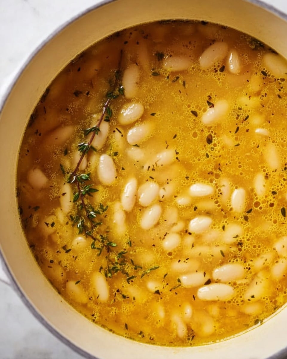 A close-up view of a white pot filled with a golden-yellow soup containing small, soft white beans floating throughout. The soup broth has a slightly oily surface with visible bubbles and small green herb flakes scattered on top. A sprig of fresh thyme rests lightly on the surface, adding texture and color variation. The pot sits on a white marbled surface, enhancing the warm colors of the soup. photo taken with an iphone --ar 4:5 --v 7