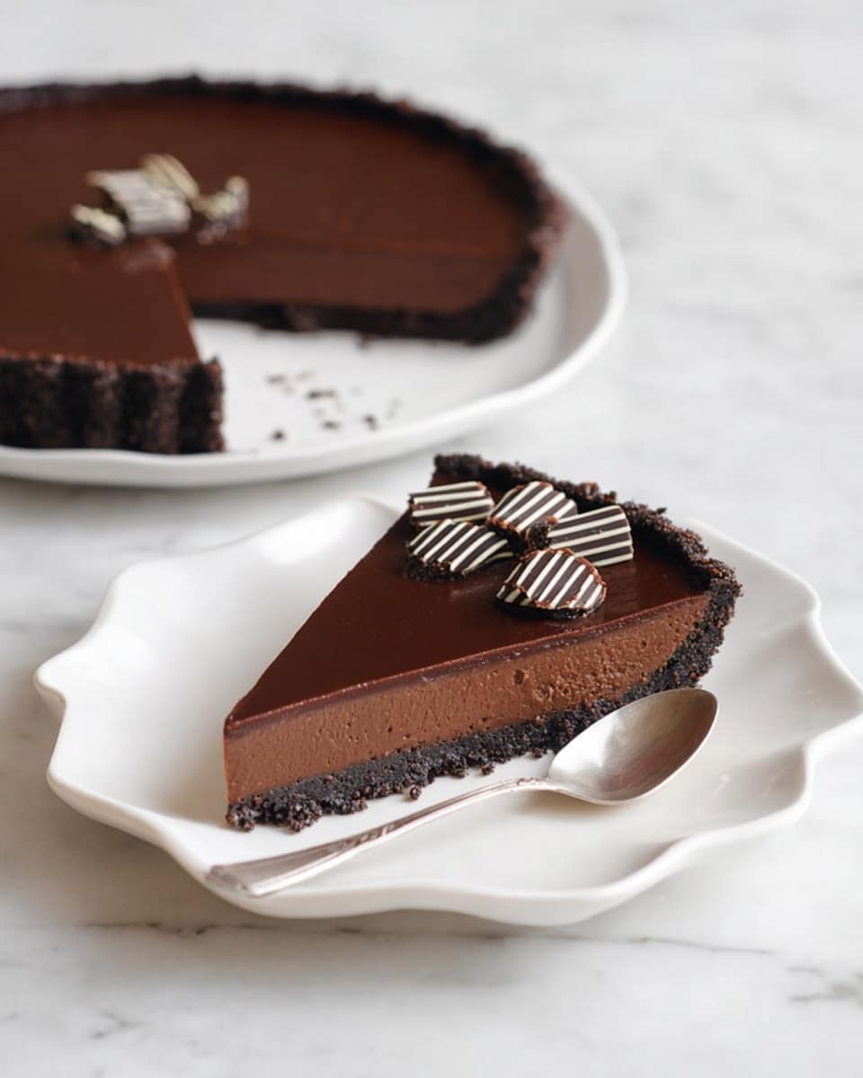 A single slice of dark chocolate tart sits on a white, wavy-edged plate on a white marbled surface. The tart has three layers: a crumbly dark chocolate crust at the bottom, a thick smooth dark chocolate filling in the middle, and a shiny dark chocolate glaze on top. The slice is decorated with several small chocolate curls that are dark and white striped, placed neatly in the center. A silver spoon lies diagonally beside the slice on the plate. In the background, the rest of the tart with one slice taken out is visible on the same white marbled surface. photo taken with an iphone --ar 4:5 --v 7