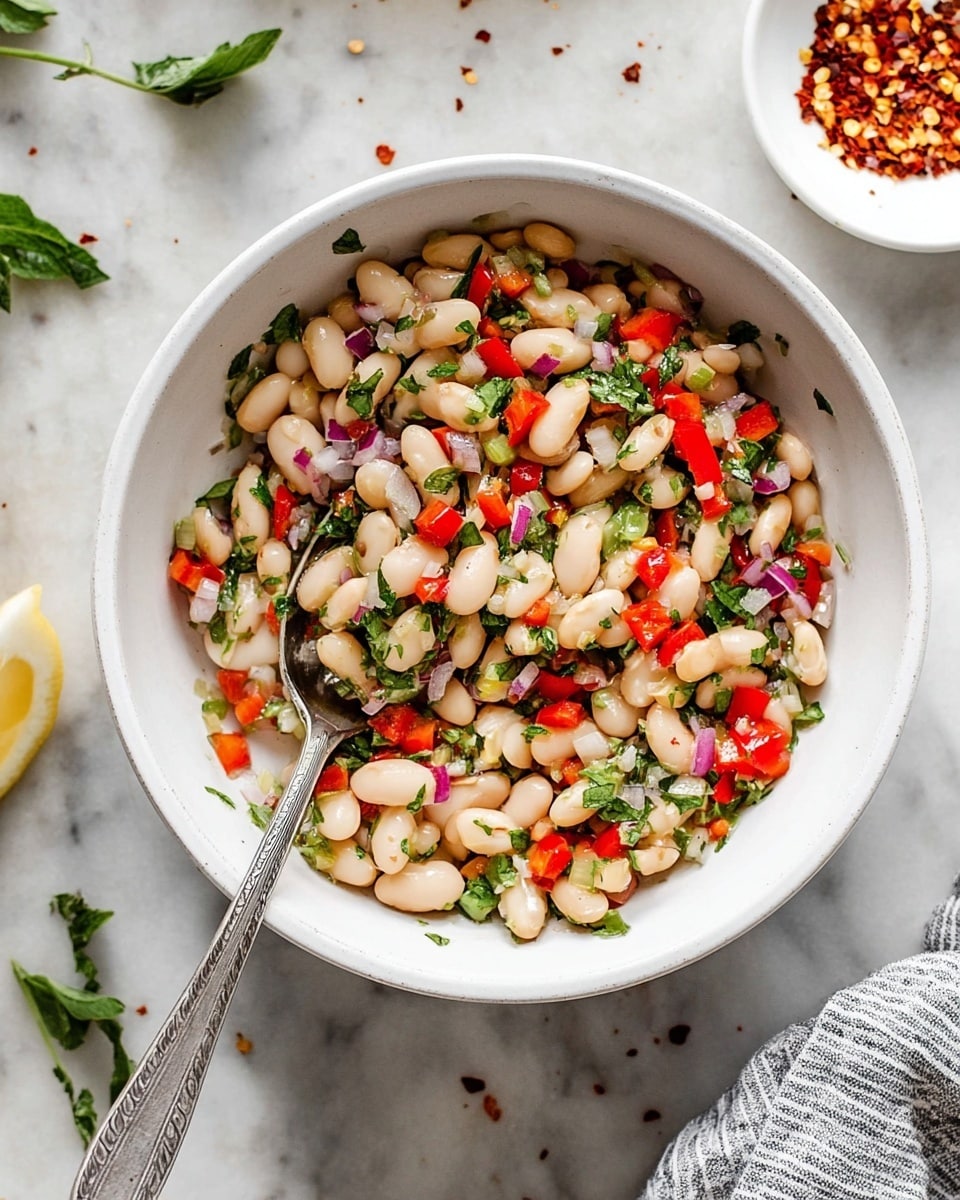A white bowl filled with a salad made of three main layers: the base layer shows white beans that are round and smooth, the middle layer contains chopped red bell peppers and small pieces of red onions adding red and purple tones, and the top layer has fresh green herbs scattered evenly. A silver spoon rests inside the bowl, partially covered by the salad. The bowl sits on a white marbled surface with a few green herb leaves around it and a white bowl with red chili flakes nearby. A piece of a striped cloth is visible at the bottom right corner. Photo taken with an iphone --ar 4:5 --v 7