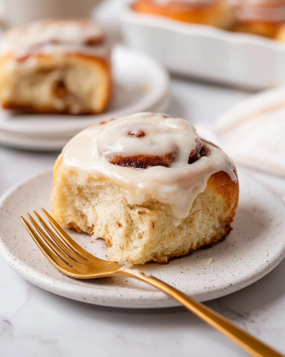 A single cinnamon roll sits on a white plate with small speckles, on a white marbled surface. The cinnamon roll has two visible layers: a soft, fluffy light golden-brown dough base with a swirl of darker cinnamon filling in the middle, topped with a thick layer of creamy white icing that drips slightly down the sides. In front of the plate lies a shiny gold fork. In the background, blurred, is another white plate with more cinnamon rolls. Photo taken with an iphone --ar 4:5 --v 7