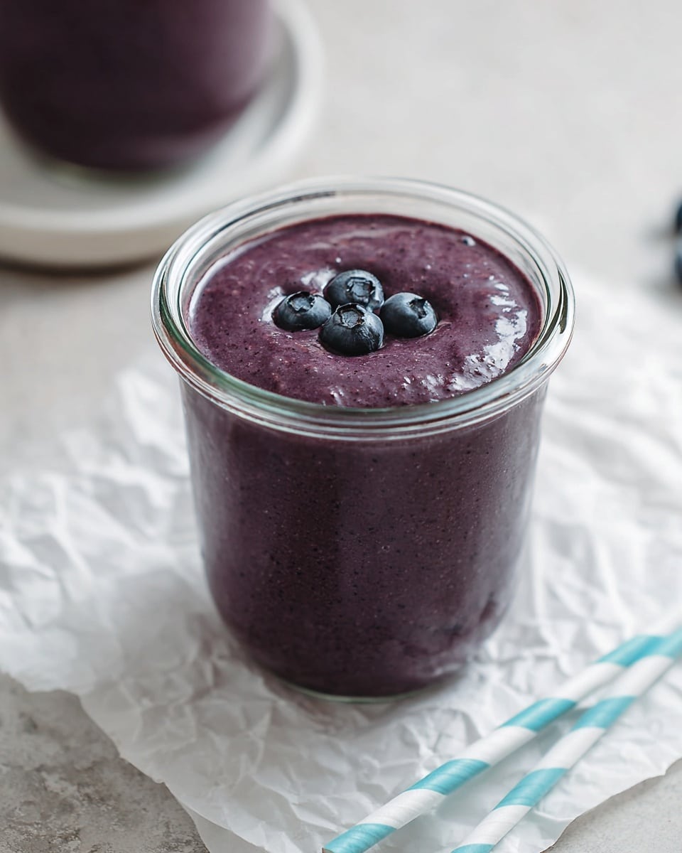 A clear glass jar filled with a smooth, thick, deep purple smoothie, topped with three whole blueberries resting gently on the surface. The jar is placed on crumpled white parchment paper set on a white marbled surface, with a striped blue and white paper straw lying beside it. Part of a second identical jar and a white plate are blurred in the background, all softly lit to highlight the rich color and texture of the smoothie. photo taken with an iphone --ar 4:5 --v 7