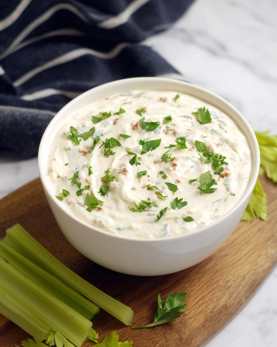 A white bowl filled with a creamy, thick white dip that has small bits of brown mixed inside, topped with small green parsley leaves scattered on the surface. The bowl sits on a wooden board with a few green celery sticks placed nearby. The background shows a white marbled surface and a dark navy towel with thin white stripes. Photo taken with an iphone --ar 4:5 --v 7