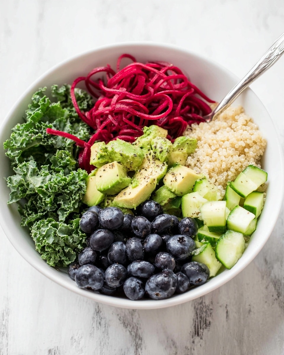 The image shows a white bowl filled with six layers of fresh ingredients arranged side by side. Starting from the left, there are dark green curly kale leaves with a bumpy texture, next to bright red spiralized beetroot strands, then light green avocado chunks sprinkled with black pepper, next to a fluffy beige layer of cooked quinoa, and at the front right there is a cluster of round, dark blue blueberries with a slight shine. In front of the kale and beetroot layers are diced cucumber pieces that are pale green with visible skin edges. The bowl sits on a white marbled surface, and a silver spoon is visible inside the bowl. photo taken with an iphone --ar 4:5 --v 7