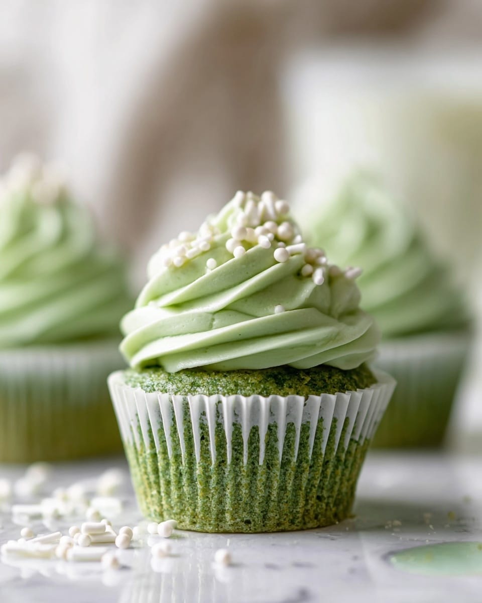 A close-up view of a green cupcake with two main layers, the bottom one is a green textured cake base in a white paper liner, and the top one is light green swirled frosting with a smooth, creamy texture, decorated with white sprinkle pearls and rods on top. The cupcake sits on a white marbled surface with some white sprinkles scattered around, and the background is softly blurred with neutral tones. Photo taken with an iphone --ar 4:5 --v 7