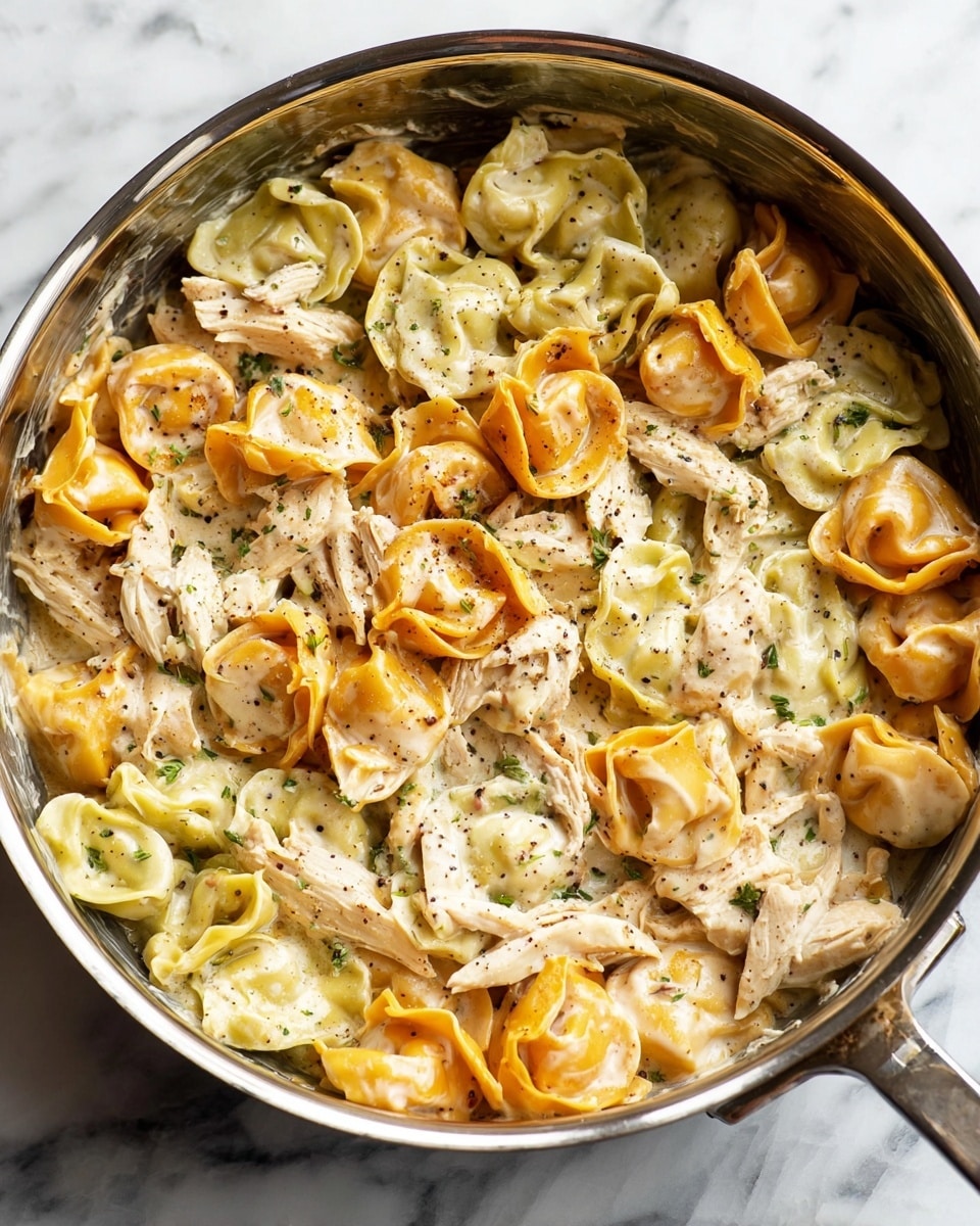 A close-up of a silver pan filled with three-color tortellini pasta layered with shredded white chicken and creamy white sauce with specks of black pepper and herbs scattered throughout. The tortellini come in three shades: orange, green, and pale yellow, creating a mixed, textured look. The chicken is spread in uneven clusters across the top and mixed into the pasta, blending into the creamy sauce that coats everything. The pan sits on a white marbled surface. photo taken with an iphone --ar 4:5 --v 7