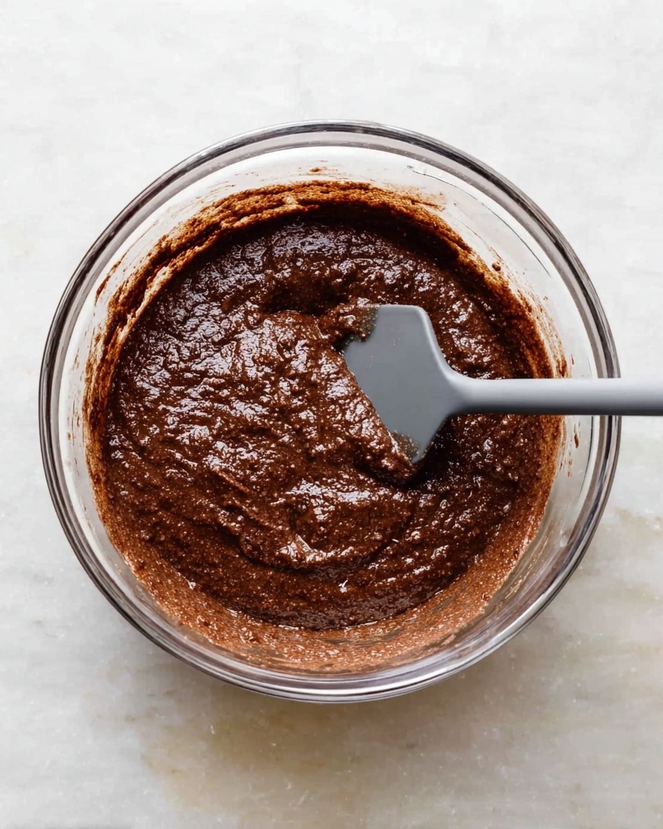 A clear glass mixing bowl is filled with thick, dark brown batter that looks slightly lumpy with a shiny, moist surface. A gray spatula is placed inside the bowl, partially mixing the batter, with some batter sticking to its edge. The bowl sits on a white marbled surface, which adds a clean and bright background to the image. photo taken with an iphone --ar 4:5 --v 7