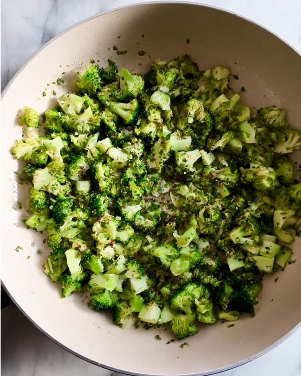 The image shows a white pan filled with chopped green broccoli pieces cooked lightly. The broccoli pieces have a mix of small florets and stems, spread evenly across the pan’s bottom. The broccoli has a fresh green color with small herbs or seasoning sprinkled on top, giving a textured look. The pan sits on a white marbled surface with some light shadows around it. Photo taken with an iphone --ar 4:5 --v 7