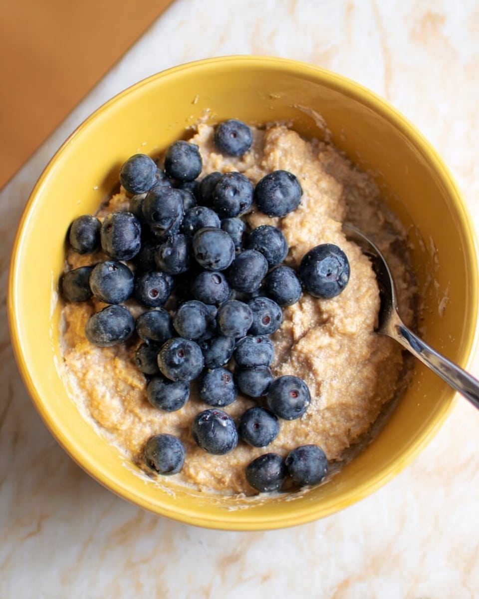 A close-up of a yellow bowl filled with thick, light brown oatmeal, topped with a cluster of fresh, round, dark blue blueberries mostly on one side, with some scattered on the oatmeal. A silver spoon is partially submerged in the oatmeal on the right side. The bowl sits on a white marbled surface. photo taken with an iphone --ar 4:5 --v 7