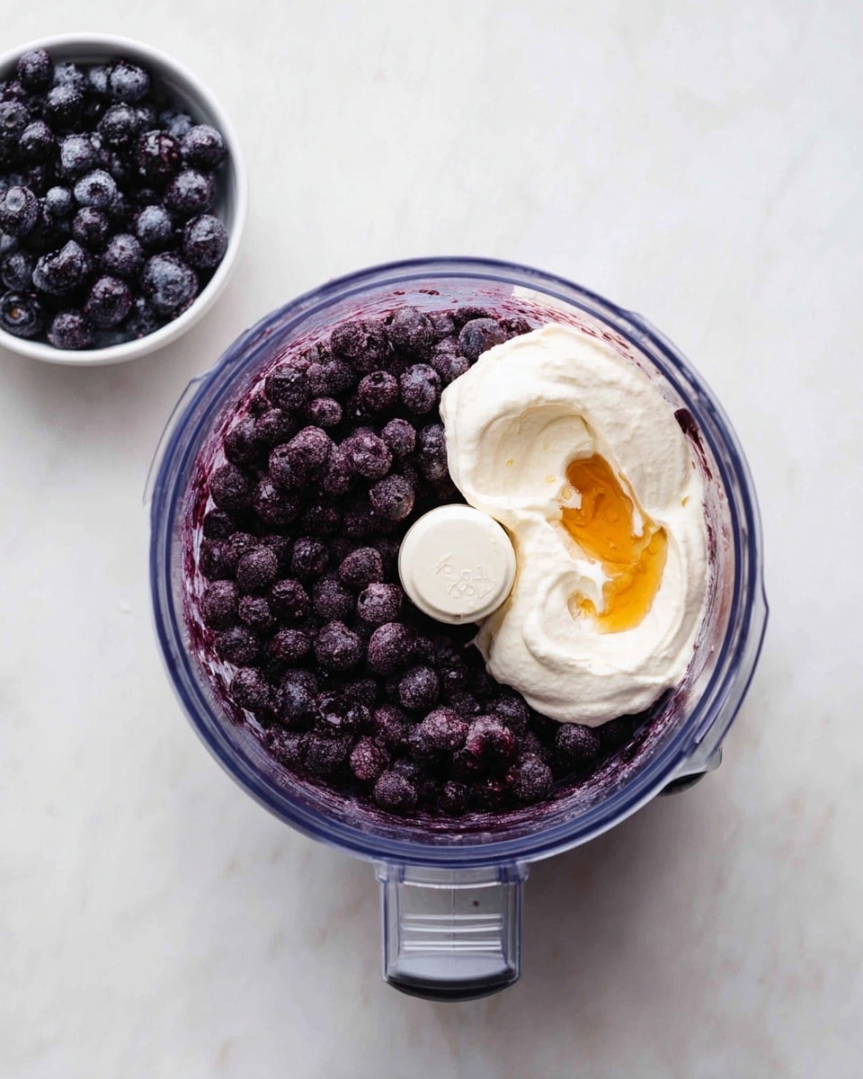 In a clear food processor bowl, there are two main layers: one side is filled with dark purple frozen berries with a matte, slightly frosted texture, and the other side holds a thick, creamy white substance with a dollop of golden syrup or honey on top. The bowl is placed on a white marbled surface, and in the top left corner, there is a small white bowl filled with the same dark purple frozen berries. The overall look is neat and arranged with a contrasting mix of deep purple, creamy white, and golden yellow. Photo taken with an iphone --ar 4:5 --v 7