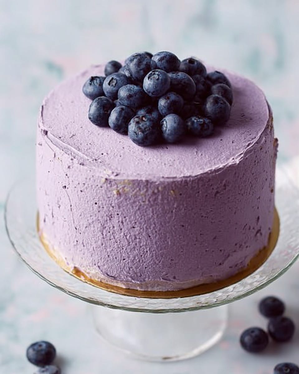 A small round cake with two visible layers covered in smooth, light purple frosting that has a slightly textured look. On top, there is a small cluster of fresh blueberries placed in the center. The cake sits on a clear glass cake stand with a thin golden trim around the edge. The background and surface beneath are white marbled textures, with a few scattered blueberries near the lower right corner. Photo taken with an iphone --ar 4:5 --v 7