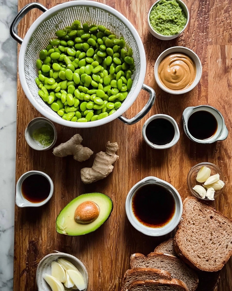 A white colander filled with bright green edamame sits on a wooden surface. Nearby, a halved avocado with smooth green flesh lies on the same surface, along with small bowls containing creamy peanut butter, fresh garlic, pieces of ginger, and some light green herb sauce. Three small cups hold different dark sauces, and a couple of slices of brown bread pop out of a white bowl in the lower right. The background is a white marbled texture. photo taken with an iphone --ar 4:5 --v 7