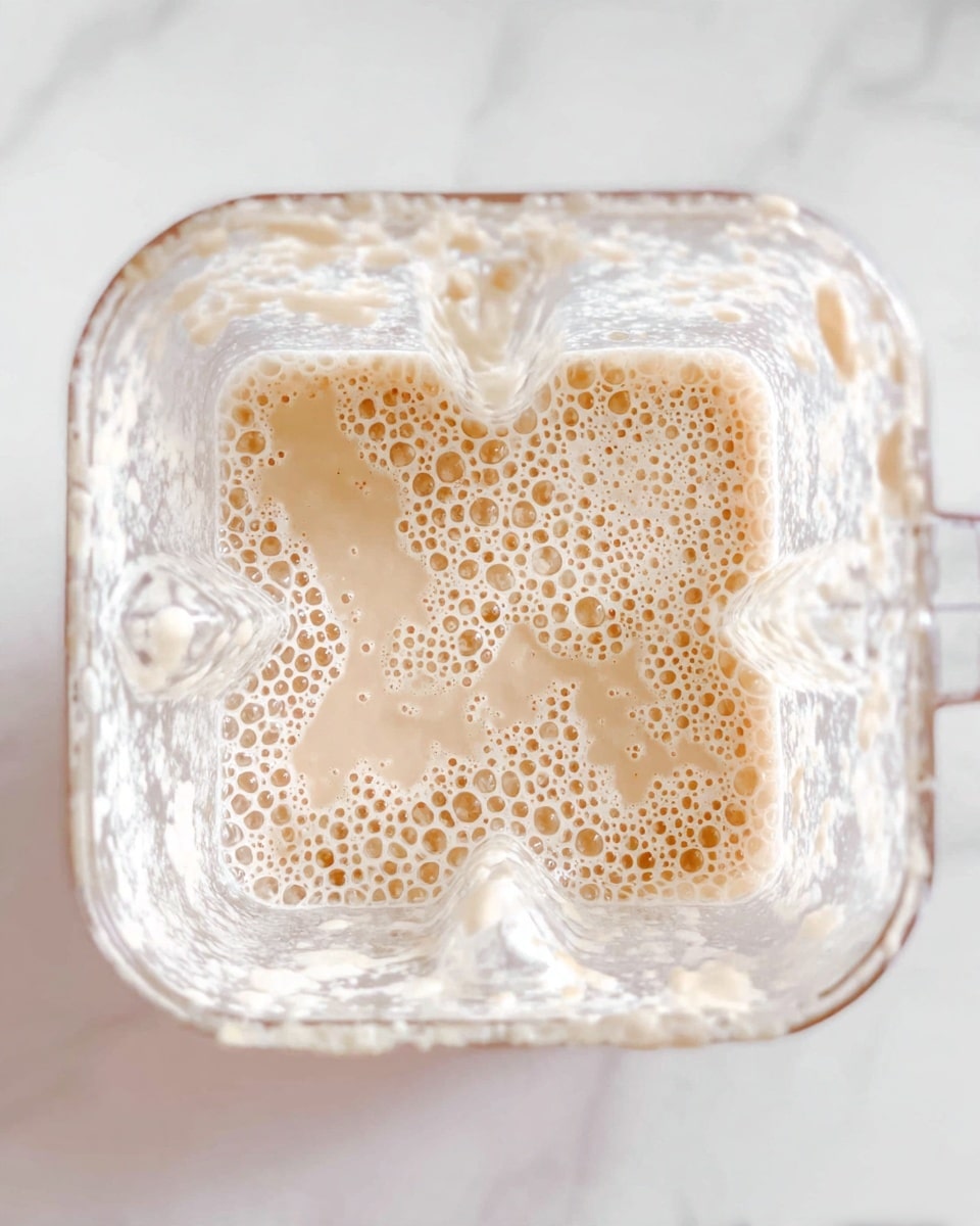 The image shows a top view of a blender container filled with a creamy, light beige liquid that has many small bubbles on its surface. The blender container is transparent with some white foam clinging to the inside sides. The background is a white marbled surface. photo taken with an iphone --ar 4:5 --v 7