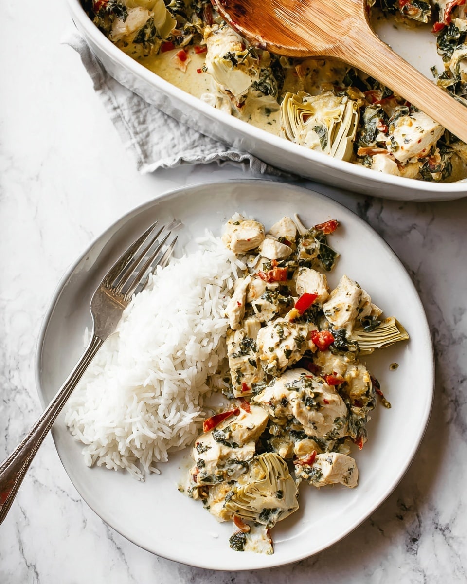 A white round plate with two main parts: on the right side, a layer of white fluffy rice with a shiny silver fork resting on it, and on the left side, a mix of chunky white chicken pieces with green leafy herbs, small red pepper bits, and artichoke hearts, all showing a creamy texture with visible spices; above, a white baking dish partially visible with similar chicken and vegetable mix, and a wooden spoon with a white handle resting on the dish, all placed on a white marbled textured surface photo taken with an iphone --ar 4:5 --v 7