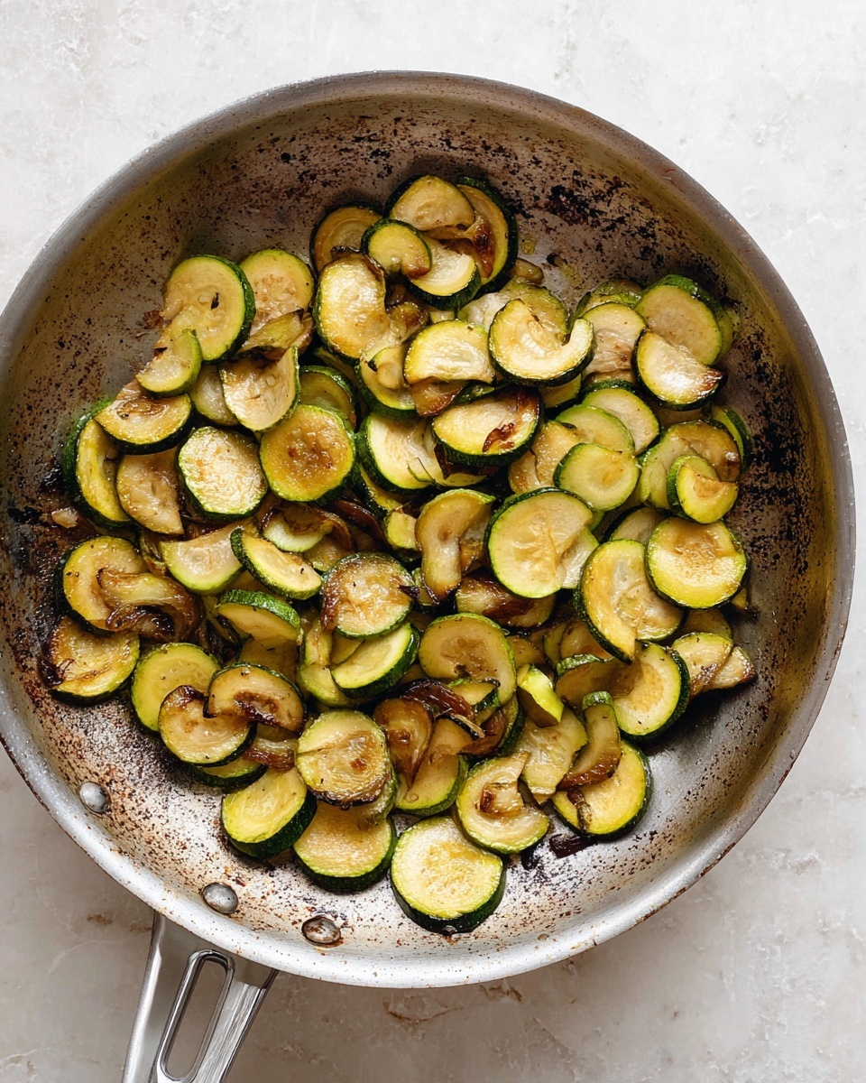 A round metal frying pan filled with cooked zucchini slices spread out in one layer. The zucchini pieces are semi-circular and irregular, showing a mix of light yellow-green soft inside and darker green skin edges, some slices are browning and caramelized, creating contrast in color and texture. The frying pan shows signs of use with dark brown and black cooking marks around the edges. The pan is placed on a white marbled textured surface, and the pan’s handle is visible in the lower part of the image. Photo taken with an iphone --ar 4:5 --v 7