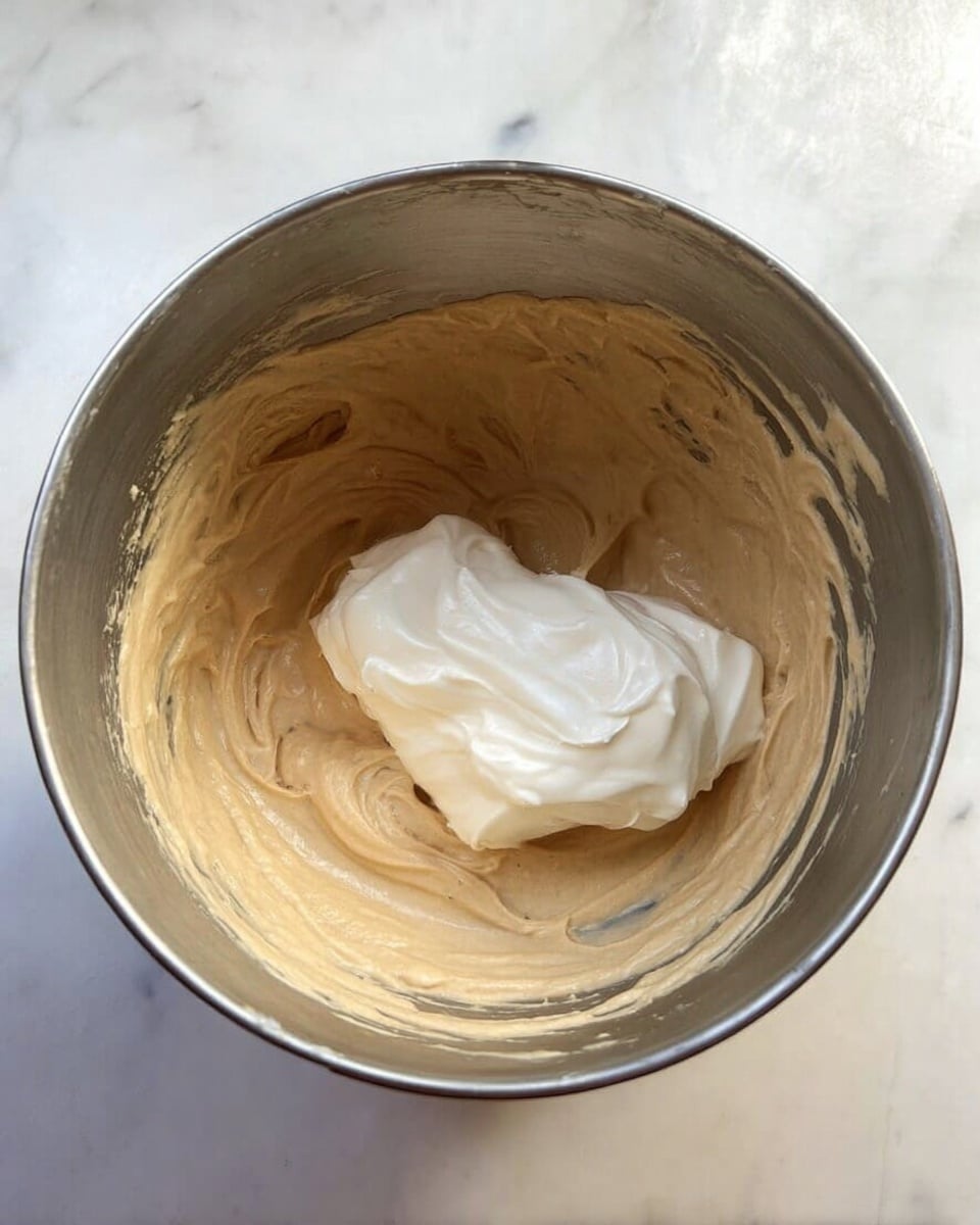 A close-up top view of a shiny metal mixing bowl with light brown batter covering the bottom and sides evenly, with a dollop of white creamy substance placed on top slightly to the right. The bowl sits on a white marbled surface. photo taken with an iphone --ar 4:5 --v 7
