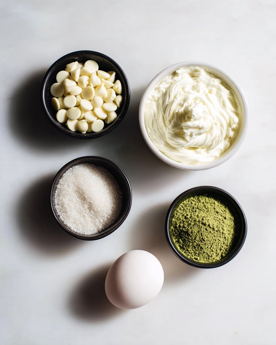 The image shows five ingredients arranged on a white marbled surface. On the top left, there is a small black bowl filled with off-white, smooth, round chocolate chips. To the right of it, there is a white bowl that holds a thick, creamy white substance with soft swirls on top. Below the chocolate chips, a small black bowl contains granulated white sugar with a slightly sparkly texture. Below the sugar and slightly to the right, there is another small black bowl containing fine bright green powder with a slightly uneven surface. On the bottom right, there is a single egg placed directly on the surface. The image is bright and well-lit with soft natural shadows photo taken with an iphone --ar 4:5 --v 7