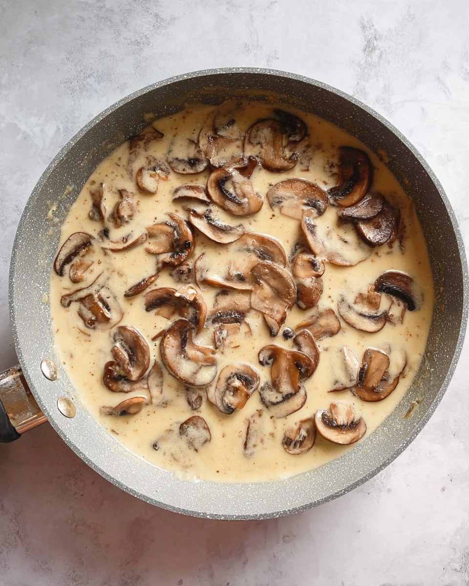 A light gray pan filled with creamy beige sauce that has many slices of brown mushrooms floating on top, the mushrooms showing their curved shapes and darker gills, all resting on a white marbled surface photo taken with an iphone --ar 4:5 --v 7