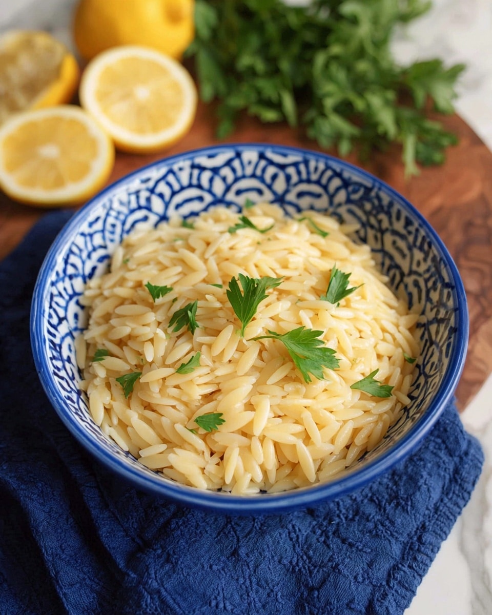 A blue and white patterned bowl filled with a single layer of small, pale yellow orzo pasta, topped with a few sprigs of fresh green parsley placed in the center. The bowl is set on a navy blue cloth over a wooden board, with bright yellow lemon wedges and green parsley blurred in the white marbled background. Photo taken with an iphone --ar 4:5 --v 7
