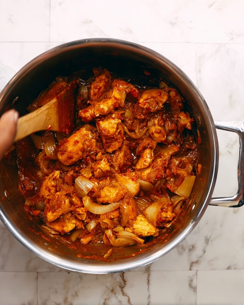 The image shows a stainless steel pot on a white marbled surface filled with pieces of chicken cooked with onions and spices. The chicken pieces are coated in a reddish-orange sauce, mixed with visible diced onions that add texture. The pot’s shiny metal handles are visible on both sides. A woman's hand holding a wooden spatula stirs the mixture, causing slightly blurred motion, which shows the sauce’s thick and rich texture. The overall look is warm and flavorful with a rustic, homemade feel. photo taken with an iphone --ar 4:5 --v 7