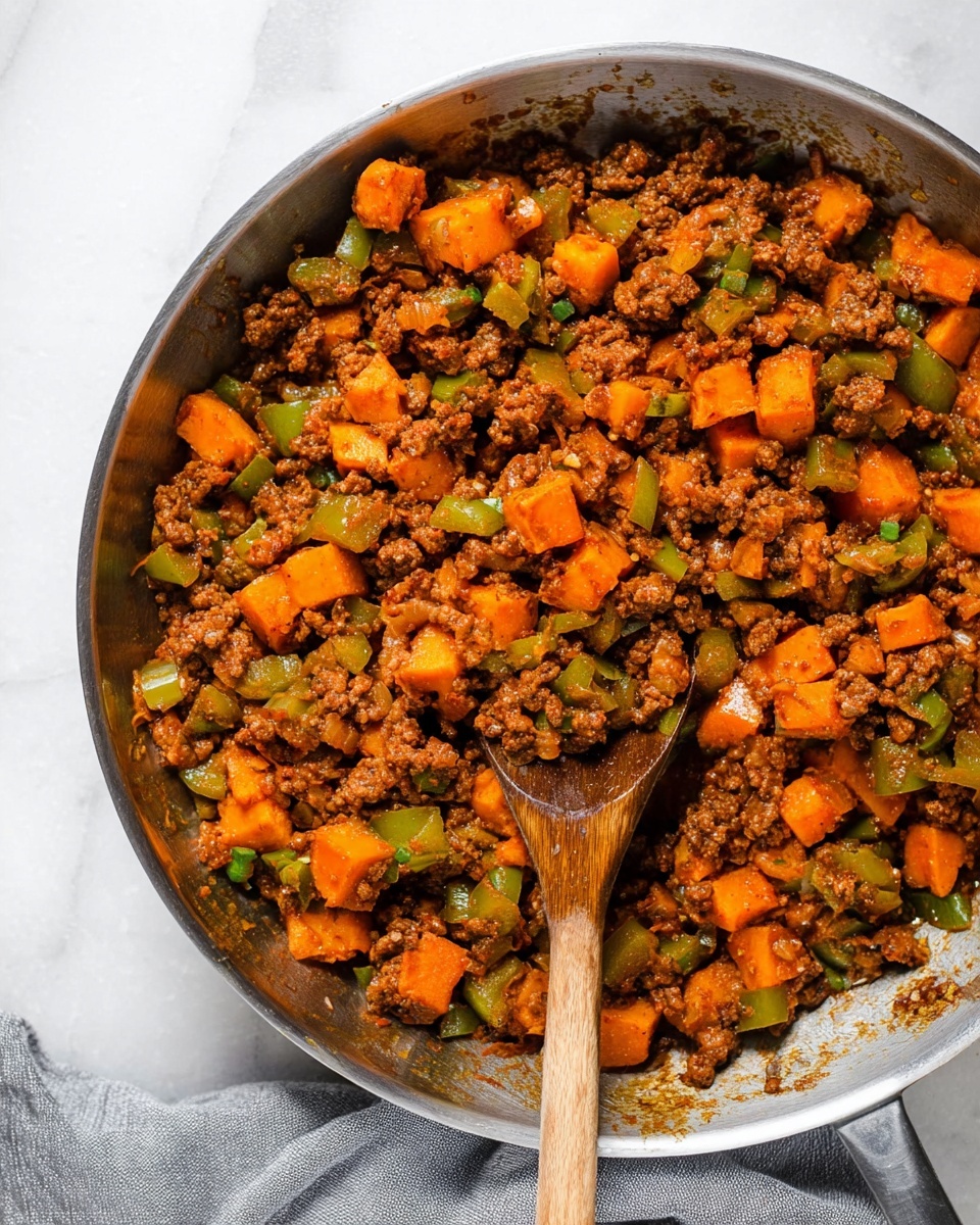 A close-up top view of cooked ground meat mixed with diced orange sweet potatoes and green bell peppers in a stainless steel pan, showing a textured mix of brown and orange chunks with hints of green, a wooden spoon resting inside the pan slightly to the left, all placed on a white marbled surface with a gray cloth partially visible at the bottom right, the dish looks juicy with some sauce visible on the pan edges and the ingredients look soft and tender photo taken with an iphone --ar 4:5 --v 7