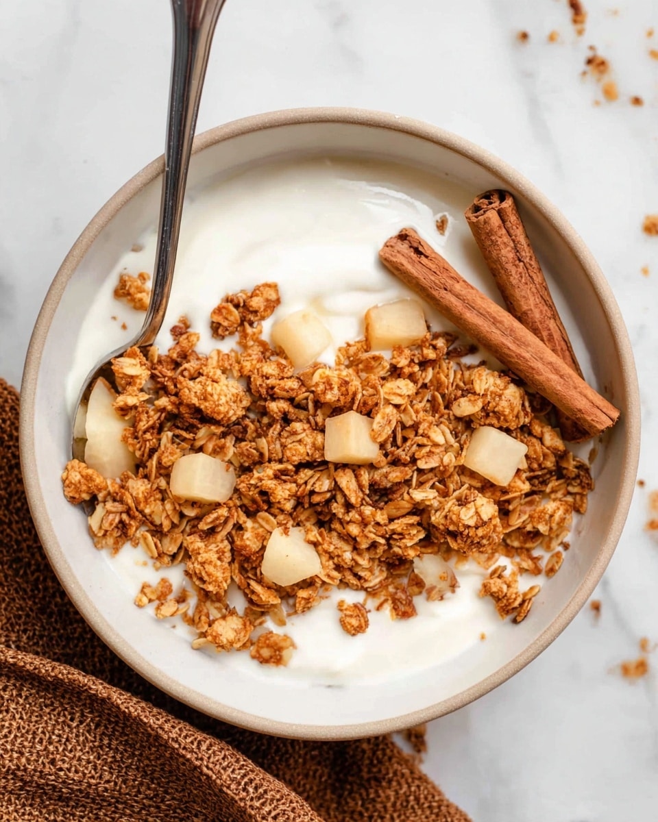 The image shows a close-up of a white bowl filled with creamy white yogurt as the bottom layer, topped with a golden brown granola mix made of oats and small clusters, and dotted with small cubes of pale yellow fruit scattered evenly on top. Two cinnamon sticks rest diagonally on the right side of the granola. A silver spoon is partially visible inside the bowl on the left side. The bowl is placed on a white marbled surface, with a brown textured cloth partially visible on the bottom right corner. photo taken with an iphone --ar 4:5 --v 7