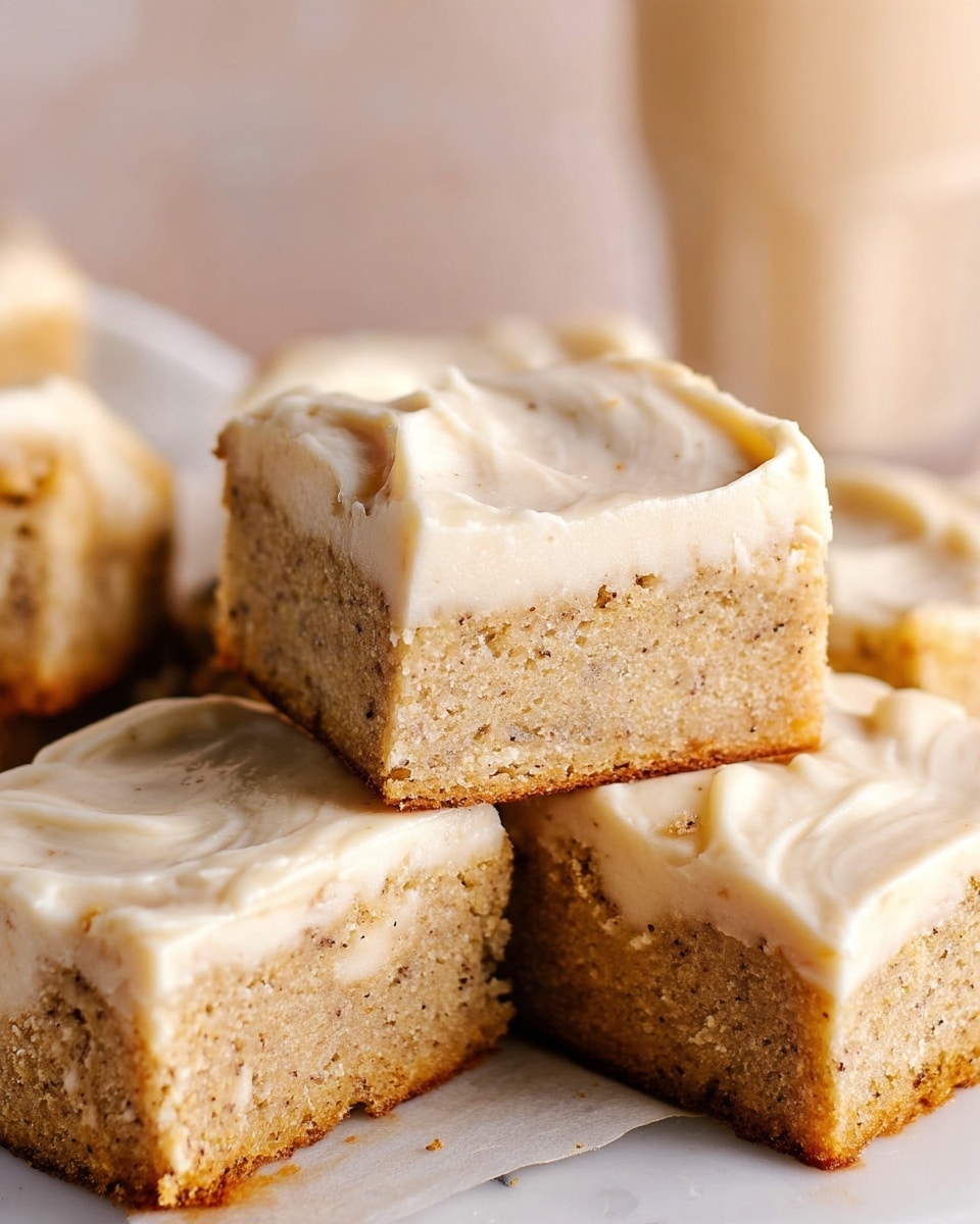 This image shows several square pieces of cake stacked closely together on a white marbled surface. Each piece has two layers: a thick, light brown cake base with a soft, moist texture filled with small specks, and a thick, creamy off-white frosting layer spread unevenly on top. The frosting looks smooth and slightly shiny with soft swirls and ridges. The background is blurred with a pale neutral color, and a glass with a light-colored drink is faintly visible. The focus is on the front cake pieces providing clear detail of the layers and texture. Photo taken with an iphone --ar 4:5 --v 7