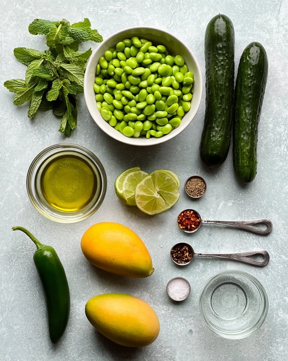 The image shows a flat lay of ingredients on a white marbled surface. At the top right, there are three small dark green cucumbers arranged horizontally. To their left, a small bunch of fresh green mint leaves rests. Below the mint, a white bowl filled with green edamame beans sits. To the right of the edamame bowl, there is a whole ripe yellow mango. Below the mango, there are two lime halves placed side by side, showing their green inside. To the right of the limes, a small clear glass bowl with a light oil is visible. On the bottom left, there is one dark green jalapeño pepper. Next to it, three metal measuring spoons hold different spices: white salt, red chili flakes, and more salt. Finally, a clear small bowl with a light liquid is positioned in the bottom right corner. photo taken with an iphone --ar 4:5 --v 7