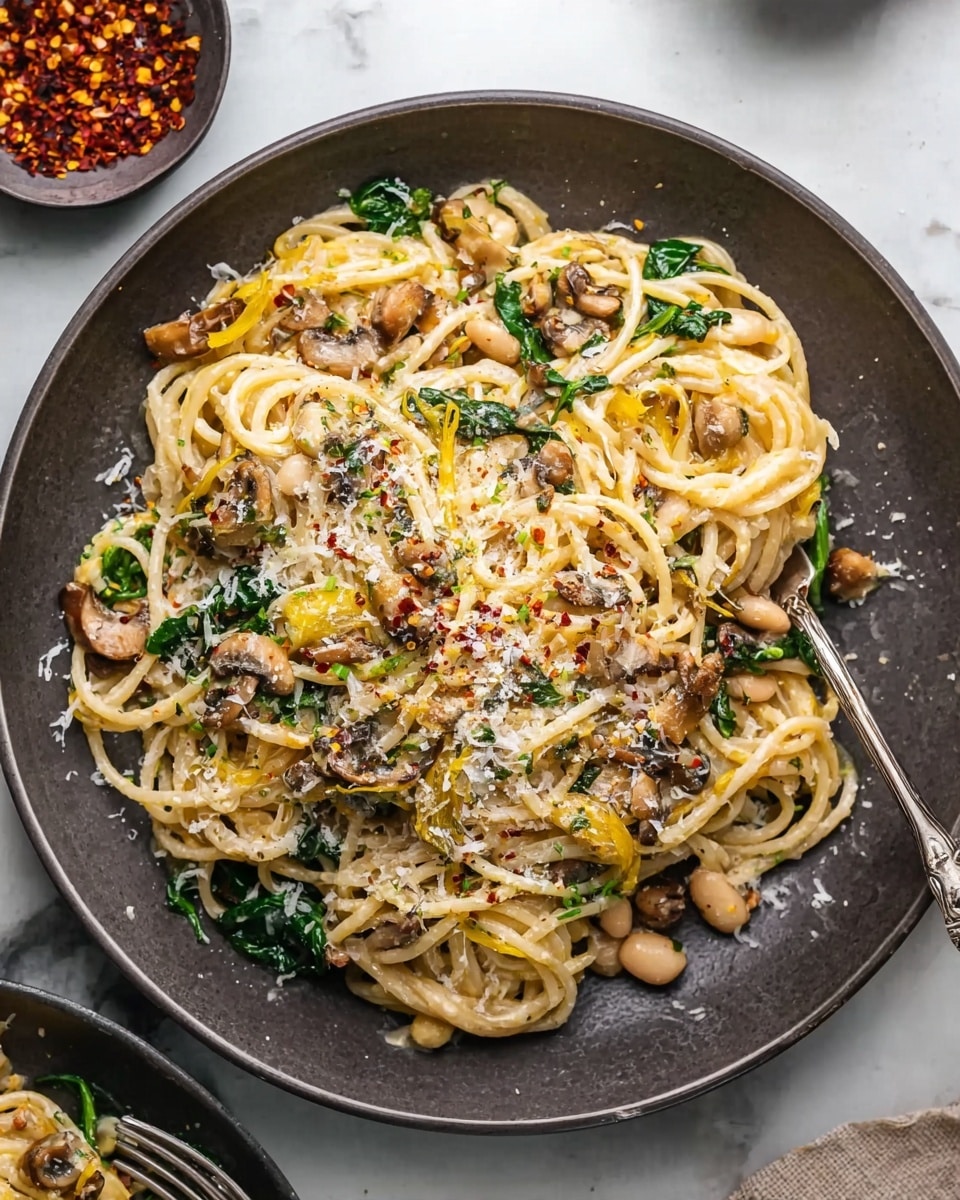 The image shows a round white plate filled with a creamy pasta dish made of spaghetti noodles mixed with sautéed mushrooms, white beans, wilted greens, and pieces of yellow squash. The top of the pasta is sprinkled with grated cheese and red chili flakes, adding texture and color contrast. The noodles are twirled into soft piles layered evenly across the plate. The background is a white marbled surface with a small bowl of red chili flakes and a fork partly in the frame near the plate. photo taken with an iphone --ar 4:5 --v 7