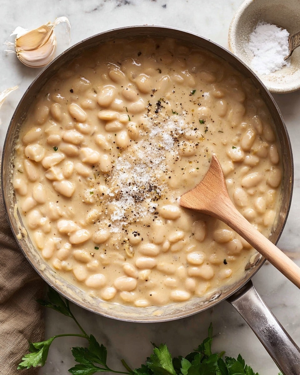 A close-up top view of a shiny metal pan filled with a thick creamy white bean stew, showing soft, light beige beans mixed in a smooth, pale sauce, sprinkled with crushed black pepper and some white seasoning in the center, with a light wooden spoon resting on the right side inside the pan. Around the pan, there is a small white bowl of coarse salt with a tiny metal spoon, a whole garlic bulb, on a white marbled surface with some green parsley leaves partially visible at the bottom and right edges. Photo taken with an iphone --ar 4:5 --v 7