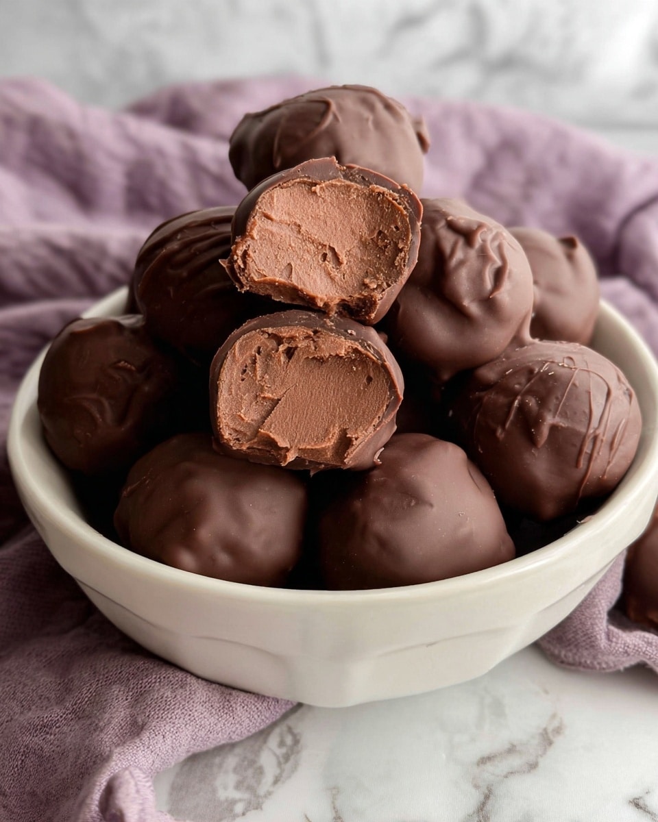 A white bowl is filled with many round chocolate-covered truffles, each with a smooth or slightly textured outer chocolate layer. One truffle is cut in half and placed on top, showing a thick, soft chocolate center inside with a rich brown color and a smooth texture. The bowl sits on a white marbled surface with a soft purple cloth in the background. The lighting is soft and natural, highlighting the glossy chocolate coating on the truffles. photo taken with an iphone --ar 4:5 --v 7