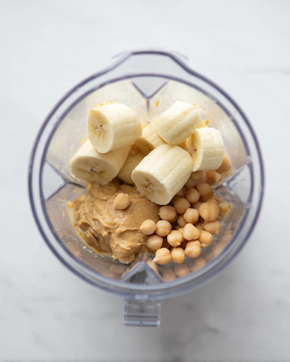 A clear blender cup is shown from above filled with six pieces of peeled bananas on top. Under the bananas, there is light brown peanut butter and a small pile of light tan chickpeas, creating three textured layers inside the blender. The background is a clean white marbled texture. photo taken with an iphone --ar 4:5 --v 7