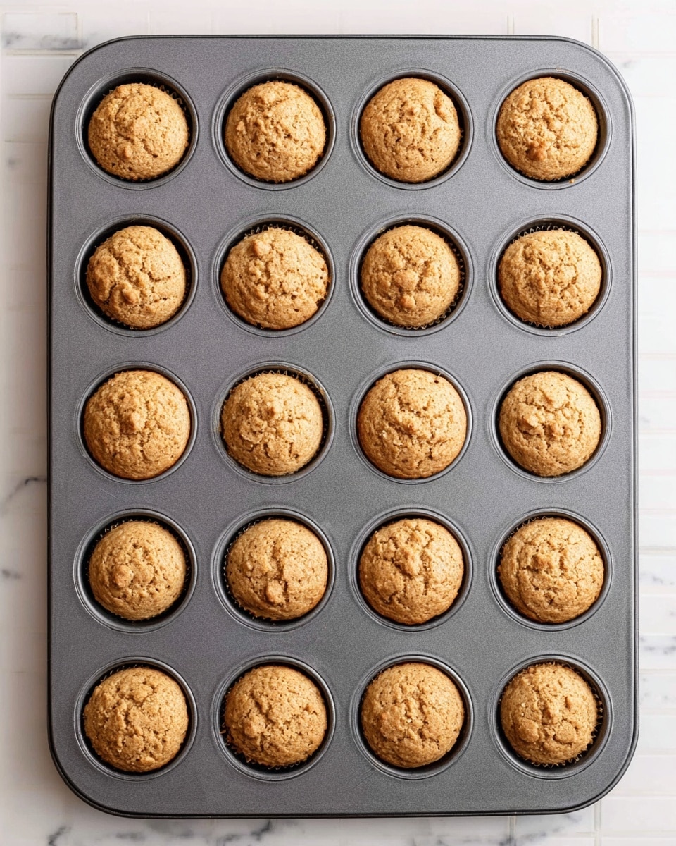 A dark gray metal baking tray with 24 round small muffins inside each mold. The muffins are light brown with slightly cracked tops and small bits visible, showing a soft texture. The tray rests on a white marbled surface with a subtle grid pattern underneath, creating contrast. The muffins are evenly placed, filling every mold, and the scene is brightly lit for a clear and fresh look photo taken with an iphone --ar 4:5 --v 7