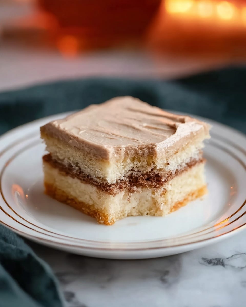 The image shows a square layered dessert on a white plate with thin gold lines. The dessert has four clear layers: the bottom layer is a pale, soft cake, the second layer is a darker, moist filling, the third layer is a light, smooth cream-like layer, and the top layer is a thick, light brown frosting with a creamy texture. A small bite is taken from one corner, showing the distinct layers inside. The plate sits on a white marbled surface, with a dark cloth underneath and a blurred, warm orange light in the background. Photo taken with an iphone --ar 4:5 --v 7