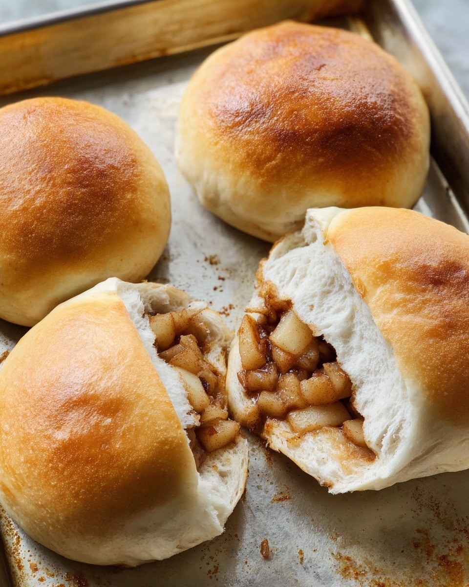 The image shows four golden brown baked buns on a metal tray, three whole and one opened to reveal a filling inside. The filling consists of small, cooked apple pieces with a soft, shiny texture, lightly coated in a cinnamon-colored sauce. The buns have a smooth, slightly shiny surface with a soft and fluffy dough visible around the edges. The opened bun is positioned diagonally on the tray, showing the apple filling packed inside the white dough layers. The tray has a slightly oily, baked texture. photo taken with an iphone --ar 4:5 --v 7
