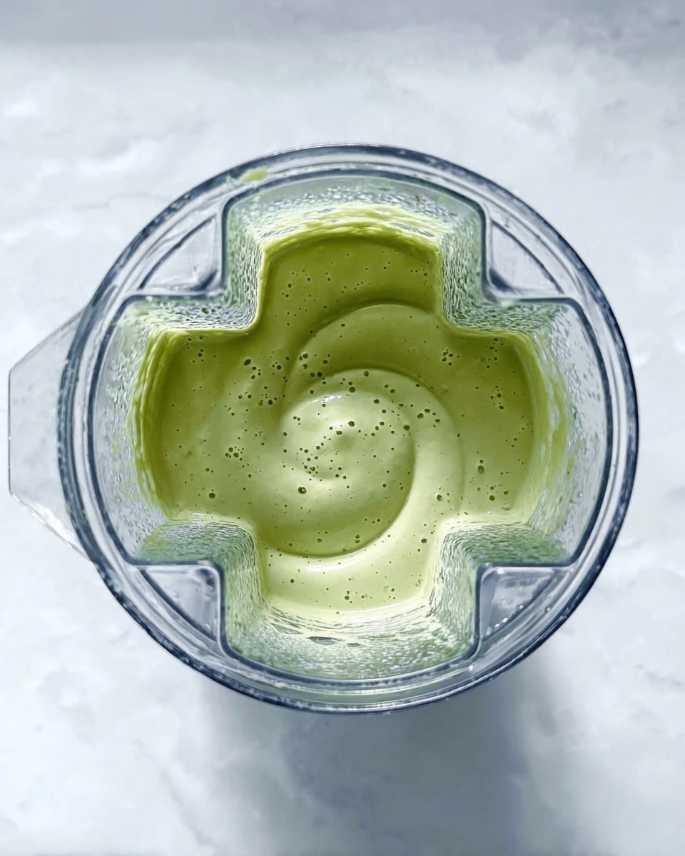 A close-up top view of a blender filled with light green creamy sauce or smoothie, showing a smooth texture with tiny bubbles on the surface, spinning in a clear blender jar with sharp diamond-like edges, placed on a white marbled texture background photo taken with an iphone --ar 4:5 --v 7