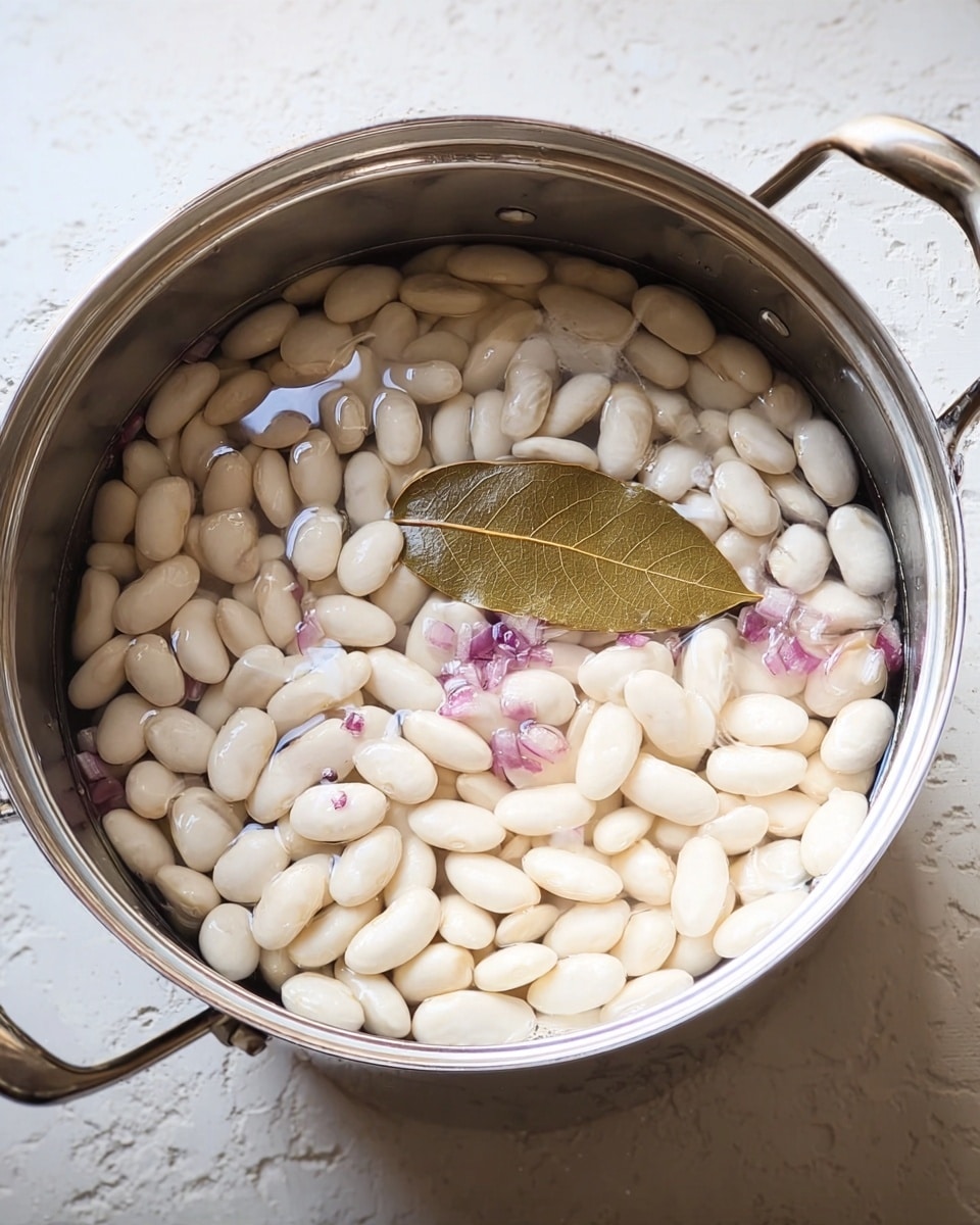 A large silver pot filled with a single layer of white beans soaking in clear water. The beans are plump, smooth, and evenly spread across the pot's bottom. In the middle, there is a brown bay leaf floating on the surface, and small bits of light purple onion pieces are scattered throughout the water. The silver pot is against a white marbled textured surface. photo taken with an iphone --ar 4:5 --v 7