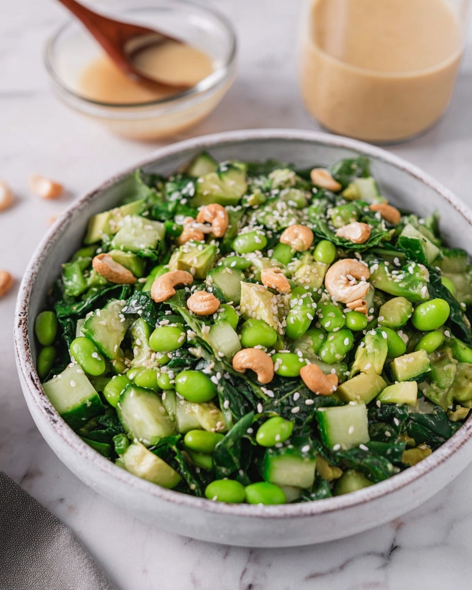 A bowl filled with a fresh salad showing layers of chopped dark green leafy vegetables and bright green edamame beans at the bottom. On top, there are light green chunks of avocado mixed with pieces of cucumber, sprinkled with white sesame seeds and small beige cashew nuts scattered throughout. The bowl is white with a slightly rough texture and rests on a white marbled surface. In the background, there is a clear glass bowl holding a light tan creamy dressing, softly blurred to keep focus on the salad. A woman's hand holding a wooden spoon is visible in the top left corner, giving a sense of action. Photo taken with an iphone --ar 4:5 --v 7