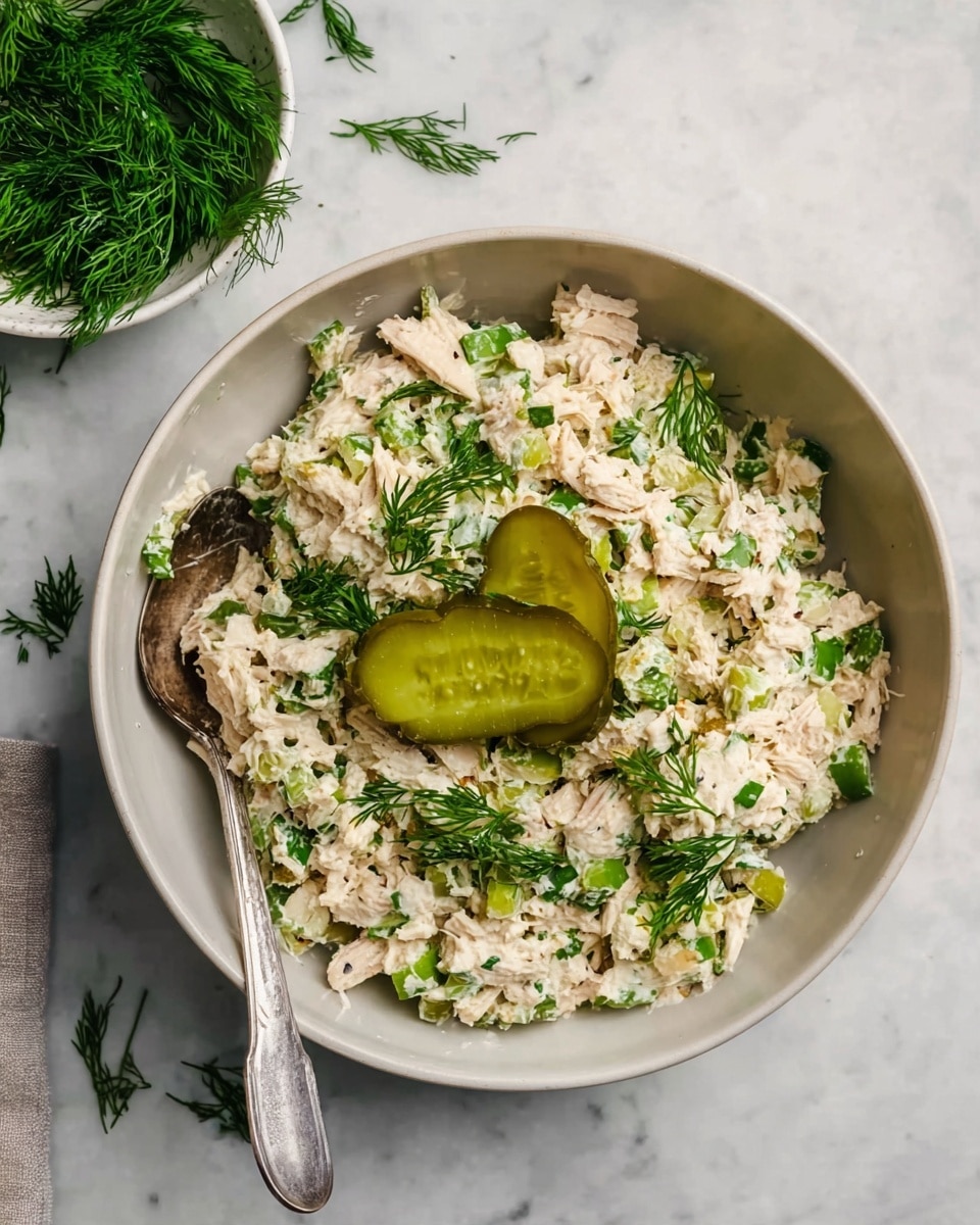 In a white bowl, there is a creamy mixture with shredded light beige pieces mixed with small green bits and specks of darker herbs, giving a textured look. On top, fresh green dill is scattered with two light green pickle slices resting in the center. A silver spoon is partly buried in the mixture at the bottom left of the bowl. The bowl sits on a white marbled surface, with some sprigs of fresh dill placed in a white bowl nearby. Photo taken with an iphone --ar 4:5 --v 7