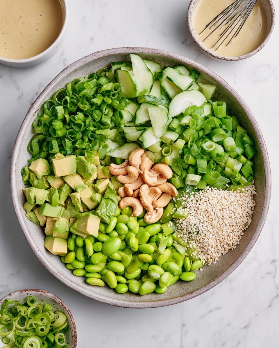 A round bowl filled with six distinct layers of fresh ingredients arranged side by side: bright green chopped cucumbers on the top right, next to chopped green onions with a shiny, leafy texture, below that light green diced avocado cubes, and bright green edamame beans with a smooth surface at the bottom. In the center, there is a small pile of light brown cashew nuts, and next to them, a sprinkle of white sesame seeds. The bowl is placed on a white marbled surface, with a bowl of light beige dressing with a whisk visible in the top right corner. Photo taken with an iphone --ar 4:5 --v 7