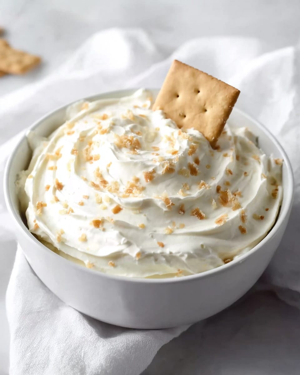 The image shows a white bowl filled with a thick, creamy white mixture topped with small light brown bits scattered evenly. A single rectangular beige cracker is placed standing upright in the middle of the creamy dip. The bowl is set on a white marbled surface with a white cloth nearby, creating a soft and clean background. The creamy mixture has swirled textures on the surface, making it look smooth and fluffy. photo taken with an iphone --ar 4:5 --v 7