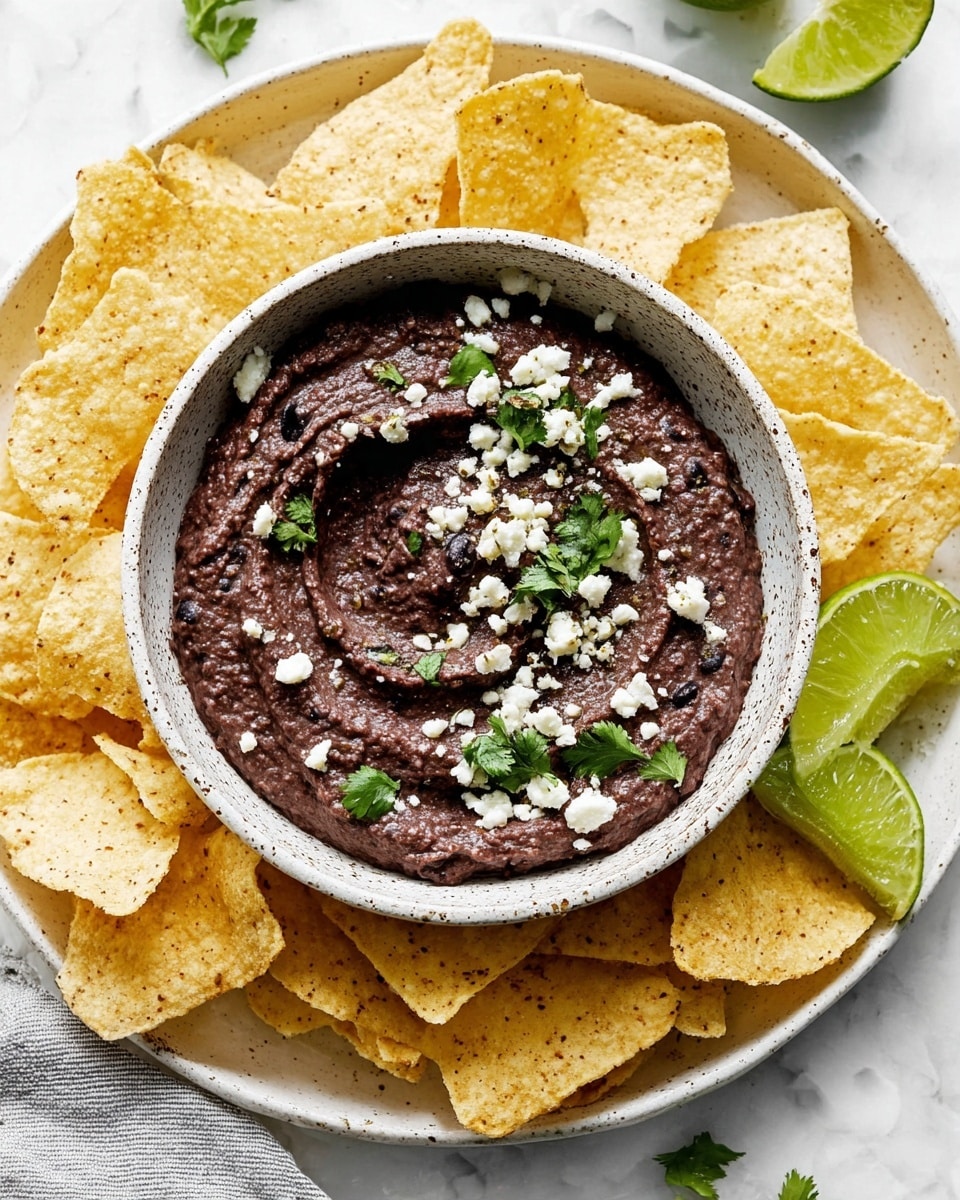 A white speckled bowl filled with dark purple black bean dip that has a thick, slightly chunky texture swirled on top. The dip is sprinkled with small white cheese crumbles and fresh green cilantro leaves. The bowl sits in the center of a large white plate, surrounded by many pale yellow triangular tortilla chips with a rough texture. On the edge of the plate are a couple of bright green lime wedges. The background has a white marbled texture. photo taken with an iphone --ar 4:5 --v 7