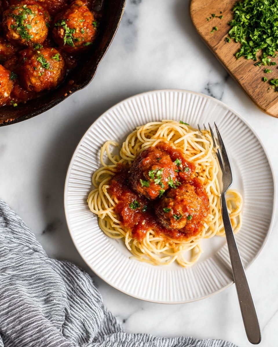 A white plate with ridged edges holds a neat mound of light brown spaghetti as the bottom layer, topped by three round meatballs coated in rich red tomato sauce. Small green parsley pieces are sprinkled over the meatballs and sauce. To the right of the plate, a silver fork rests against the edge. The plate is set on a larger plain white plate, both on a white marbled surface. In the top right, a wooden board with chopped green parsley adds color, and a cast-iron skillet with more meatballs and sauce is partially visible on the top left. A gray and white striped cloth is visible in the bottom right corner. photo taken with an iphone --ar 4:5 --v 7