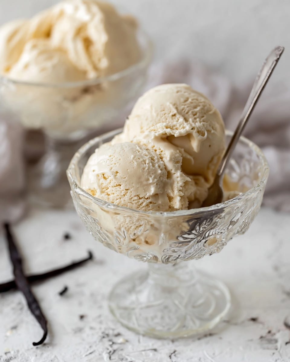 Two scoops of light beige ice cream with a creamy texture sit inside a clear, ornate glass dessert cup with a short stem. A silver spoon is placed inside the cup behind the ice cream scoops. The background shows another similar glass cup filled with the same ice cream, slightly blurred. The surface beneath is a white marbled texture with some dark vanilla pods scattered around. Photo taken with an iphone --ar 4:5 --v 7