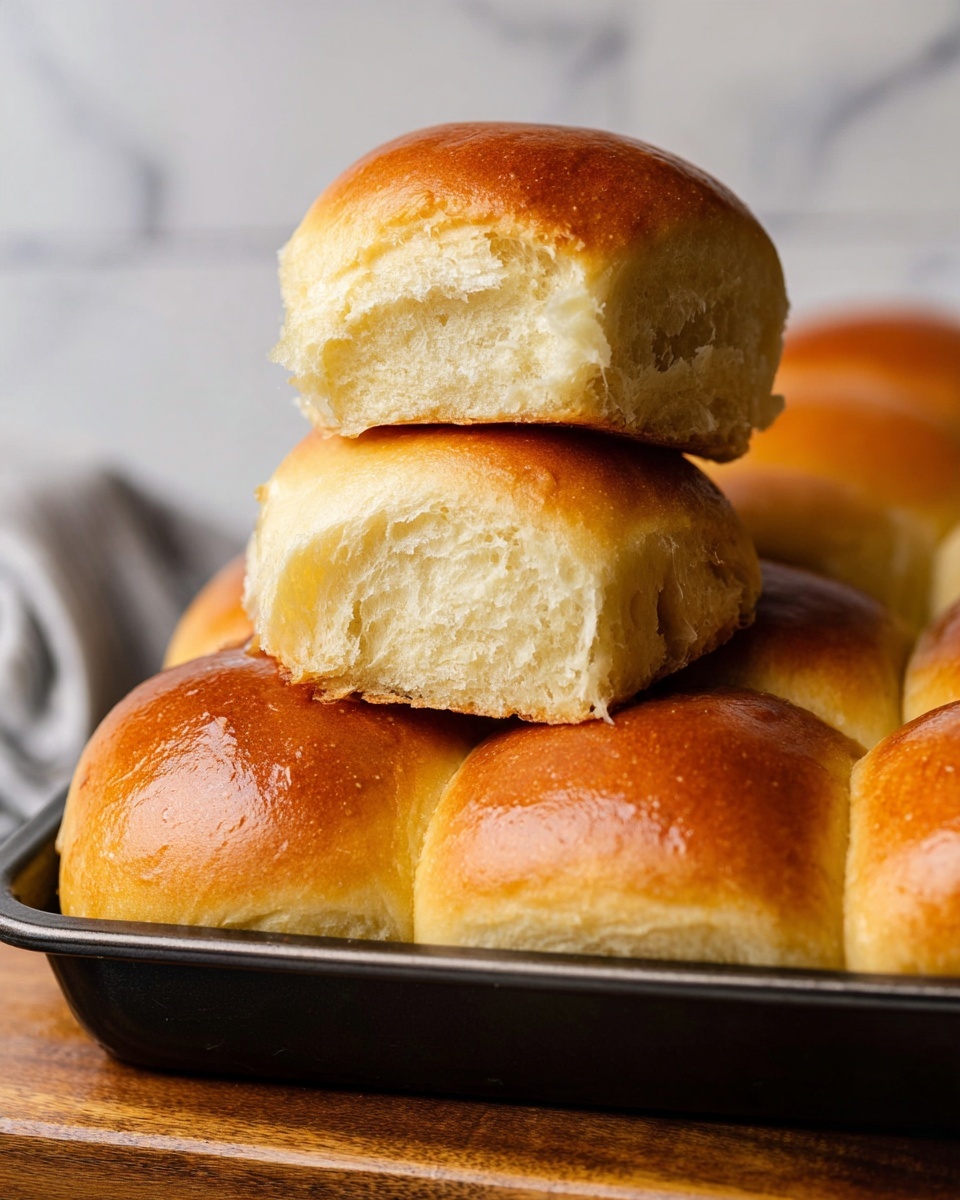 The image shows a close-up of soft, golden brown dinner rolls stacked in two layers on a dark baking tray. The bottom layer is made up of several shiny, smooth rolls with a warm brown crust, while the top layer features two rolls placed one on top of the other, both with a golden crust and a fluffy, light beige inside visible where the rolls have been gently pulled apart. The rolls have a soft and airy texture with a slight sheen on the surface, suggesting they are fresh and warm. The tray rests on a wooden surface with a white marbled background behind. Photo taken with an iphone --ar 4:5 --v 7