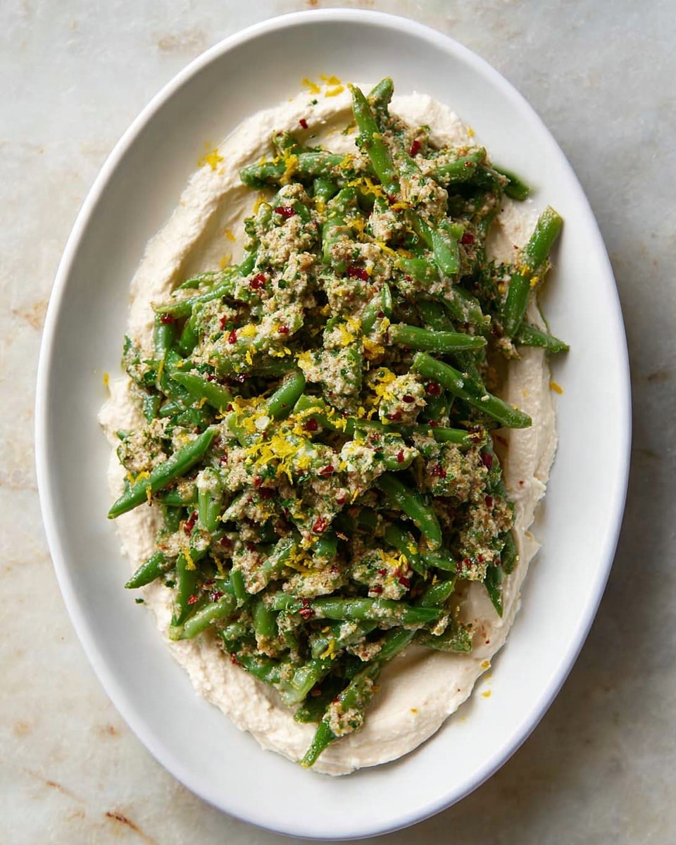 A white oval plate sits on a white marbled surface, filled with two visible layers of food. The bottom layer is a smooth, creamy white spread covering the plate in an uneven circle with slight texture on the edges. On top of this is a generous heap of chopped green beans mixed in a creamy beige sauce, showing small pieces of red chili flakes and a rough texture. Tiny yellow lemon zest bits are sprinkled over both the beans and spread, adding a bright contrast to the dish. The overall look is fresh with a mix of soft and chunky textures. photo taken with an iphone --ar 4:5 --v 7
